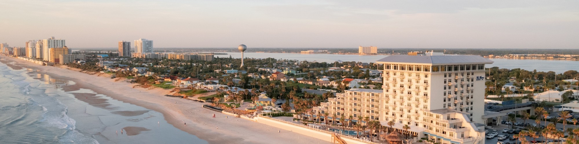 A coastal beach scene shows sandy shores, ocean waves, and a cityscape with several buildings under a clear sky.