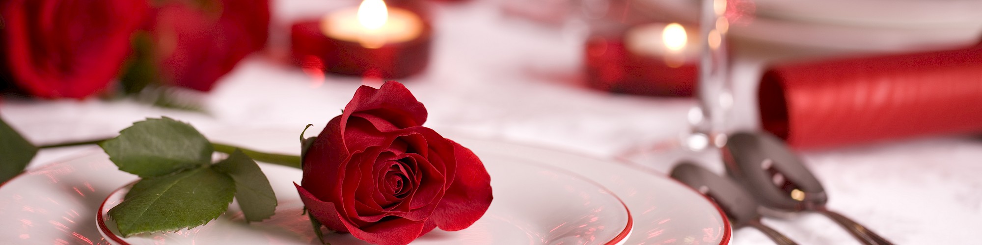 A romantic table setting with a red rose on a plate, red glassware, candles, and a bokeh effect in the background.