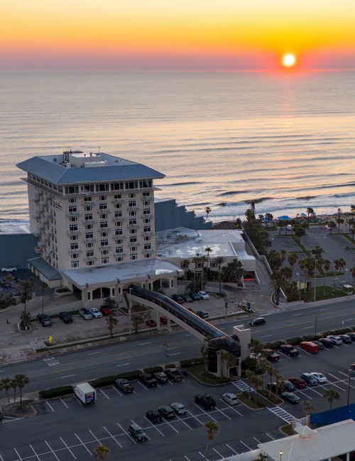 A beachfront hotel at sunset, with parking lots and a road in front, overlooking the ocean waves and a clear sky.