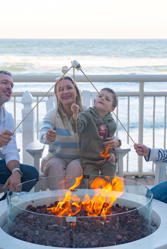 A family sits around a lit fire pit on a beach deck, laughing and roasting marshmallows together by the waves at sunset.