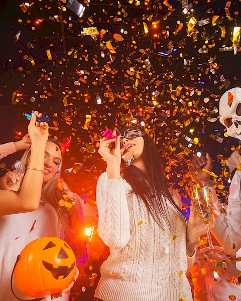 People celebrating Halloween at a party with costumes, masks, a pumpkin basket, and falling golden confetti.