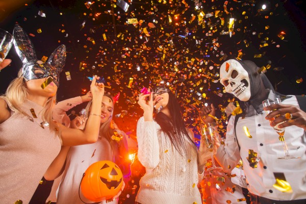 People celebrating Halloween with costumes, masks, confetti, and drinks in a festive party scene.
