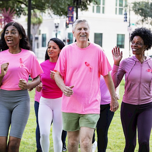 People of diverse backgrounds participate in a breast cancer awareness walk wearing pink shirts and ribbons, smiling and moving together outdoors.