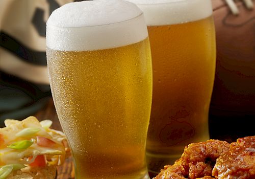 Two glasses of beer, wings, celery, and a football in the background.