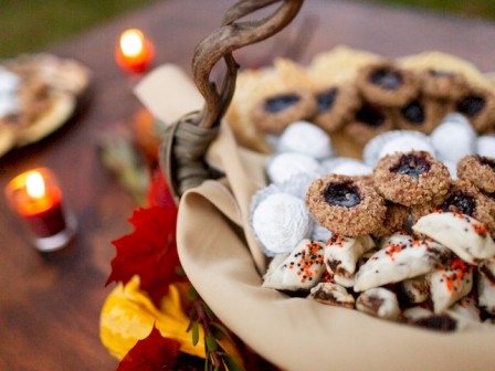 A basket of assorted cookies, powdered sugar confections, and Halloween treats on a table with candles and fall leaves.
