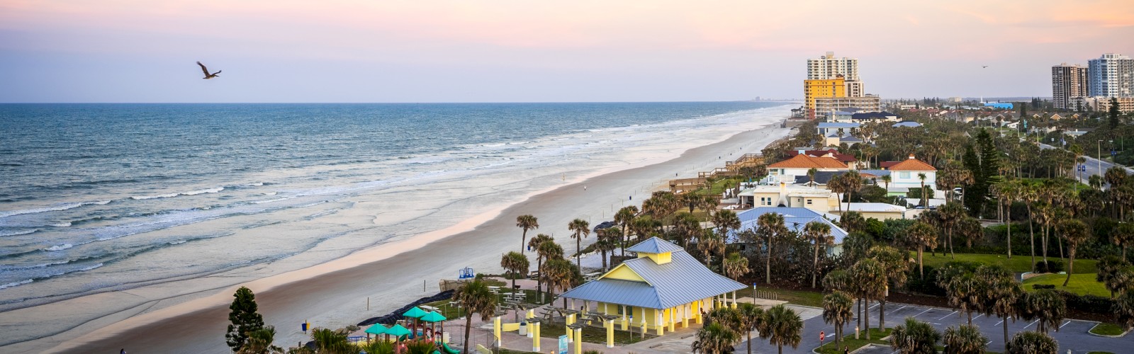A scenic view of a beach with waves, palm trees, colorful playground, and buildings in the background at sunset.