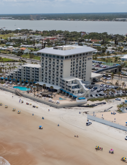 A beachfront hotel with a pool, tennis courts, and a parking lot, overlooking a sandy beach and calm ocean waters.