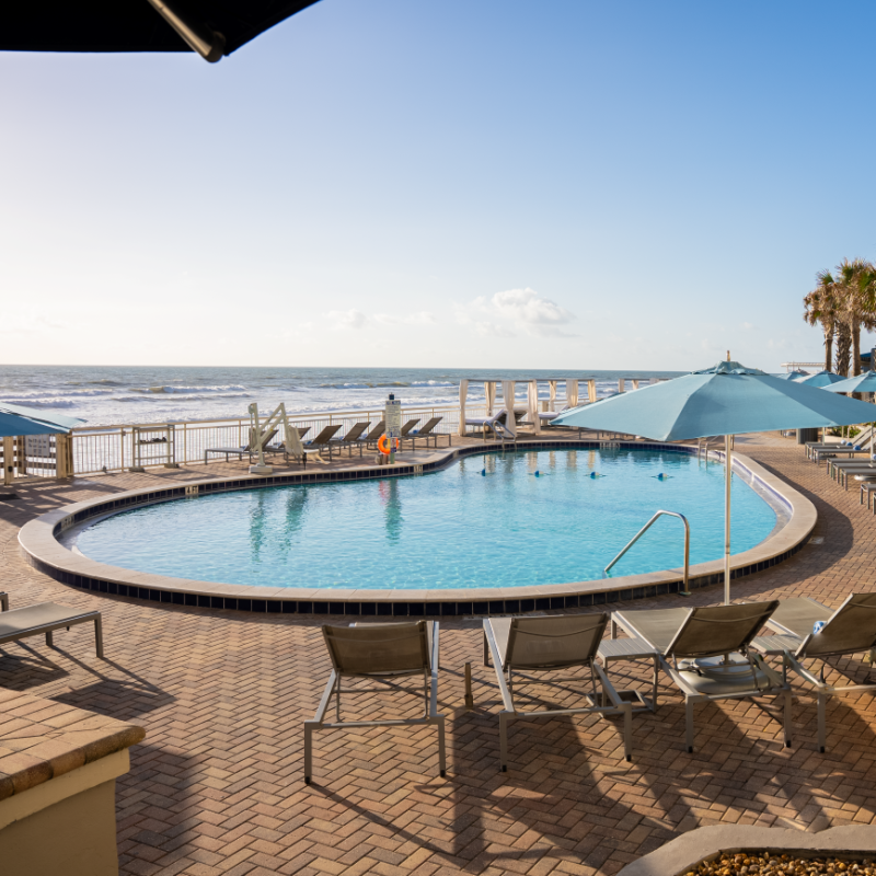 A poolside view at a beachfront hotel with lounge chairs, umbrellas, palm trees, and the ocean in the background.