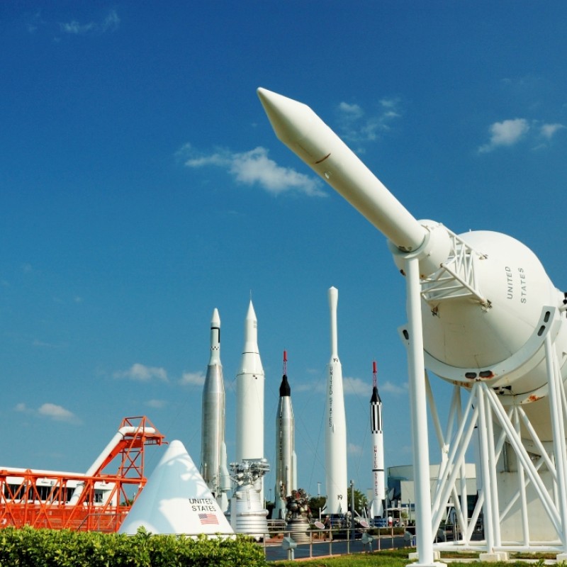 This image shows multiple rockets and space equipment displayed outdoors under a bright blue sky at a space museum or exhibit.