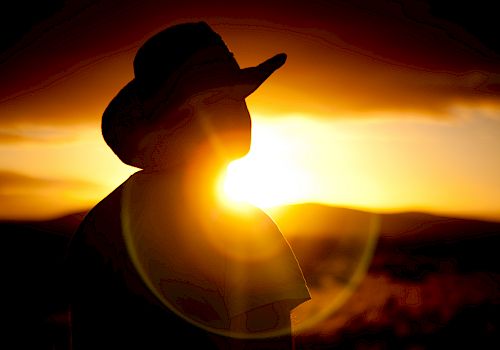 A silhouette of a person wearing a hat against a vibrant sunset background, with the sun creating a strong lens flare effect.