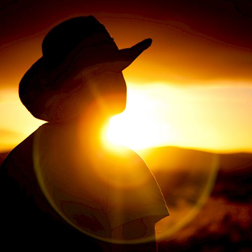 A silhouette of a person wearing a hat against a vibrant sunset background, with the sun creating a strong lens flare effect.