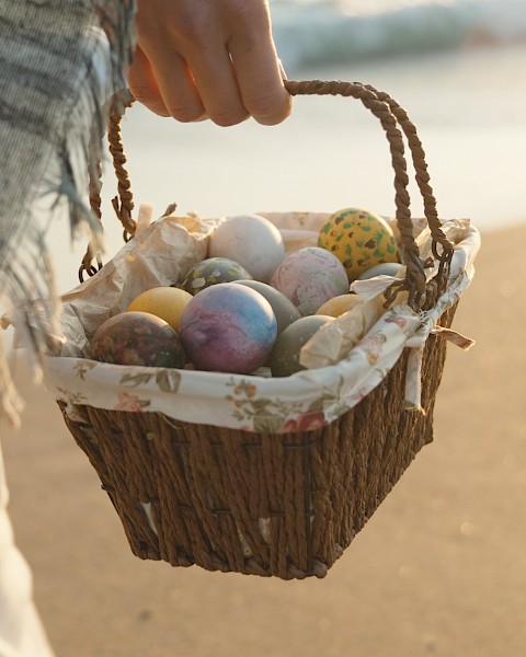 A person carries a wicker basket filled with colorful Easter eggs on a sandy beach, wrapped in a plaid blanket.