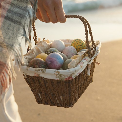 A person carries a wicker basket filled with colorful Easter eggs on a sandy beach, wrapped in a plaid blanket.