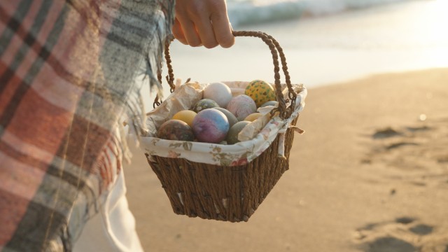 A person carries a wicker basket filled with colorful Easter eggs on a sandy beach, wrapped in a plaid blanket.