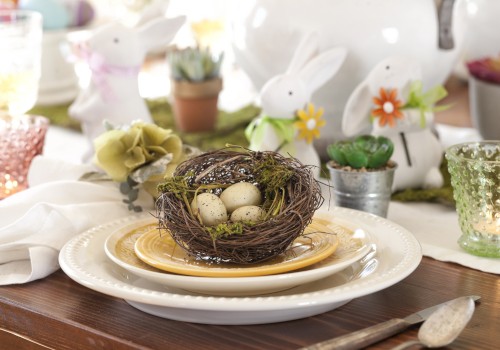 A decorative table setup with a bird's nest centerpiece containing eggs, surrounded by bunny figurines and candles, creating a spring theme.