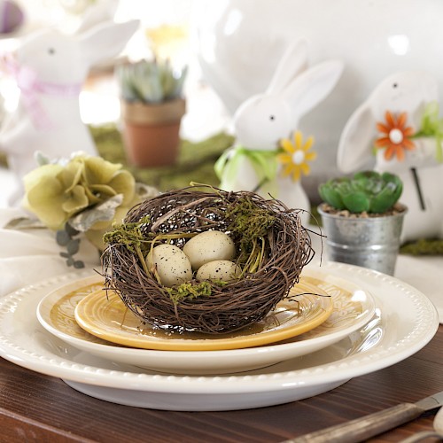 A decorative table setup with a bird's nest centerpiece containing eggs, surrounded by bunny figurines and candles, creating a spring theme.