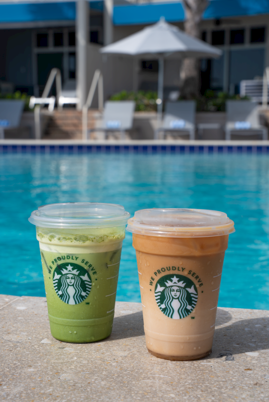 Two Starbucks drinks sit by a pool on a sunlit day, with swim chairs, an umbrella, and trees in the background.