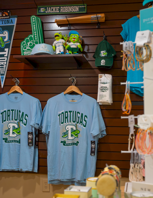 The image shows a store display with sports merchandise, including shirts, hats, plush toys, and pennants related to the Daytona Tortugas baseball team.