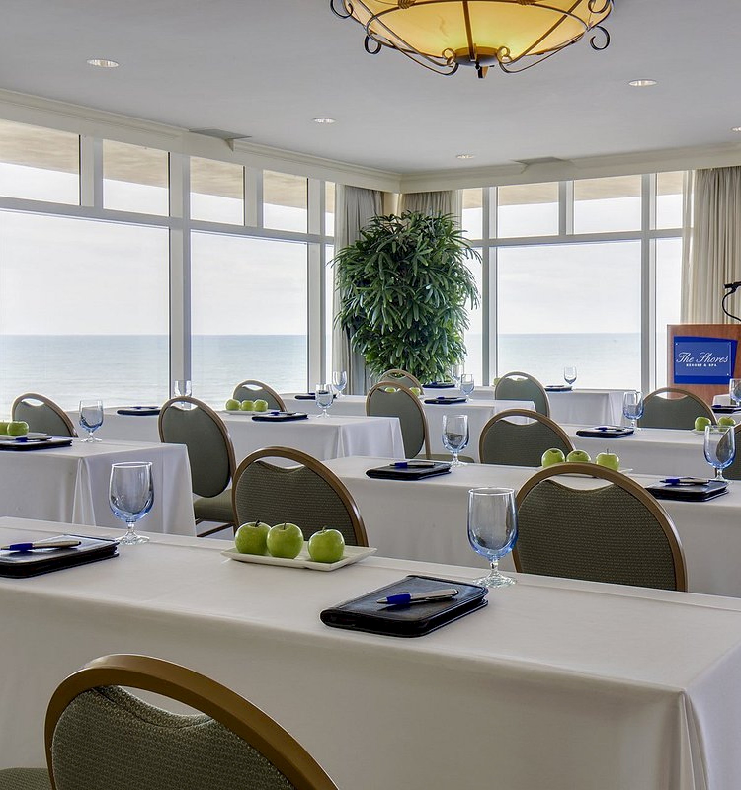 A bright, elegant conference room with sea views, set with white tablecloths, green apples, glasses, and notepads for a meeting or event.