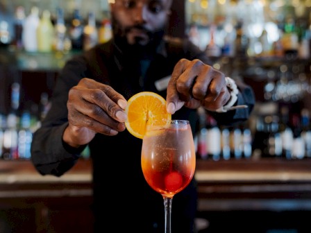 A bartender garnishes a cocktail with an orange slice in a bar setting with blurred shelves of bottles in the background.
