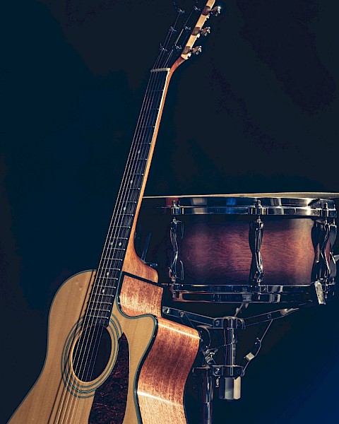 The image shows an acoustic guitar and a snare drum standing against a dark background. The guitar is leaning on the drum.