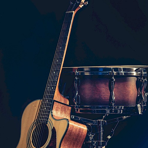 The image shows an acoustic guitar and a snare drum standing against a dark background. The guitar is leaning on the drum.