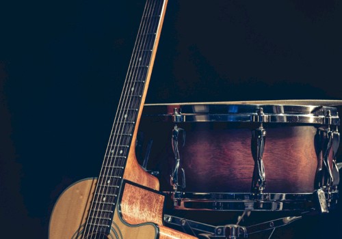 The image shows an acoustic guitar and a snare drum standing against a dark background. The guitar is leaning on the drum.