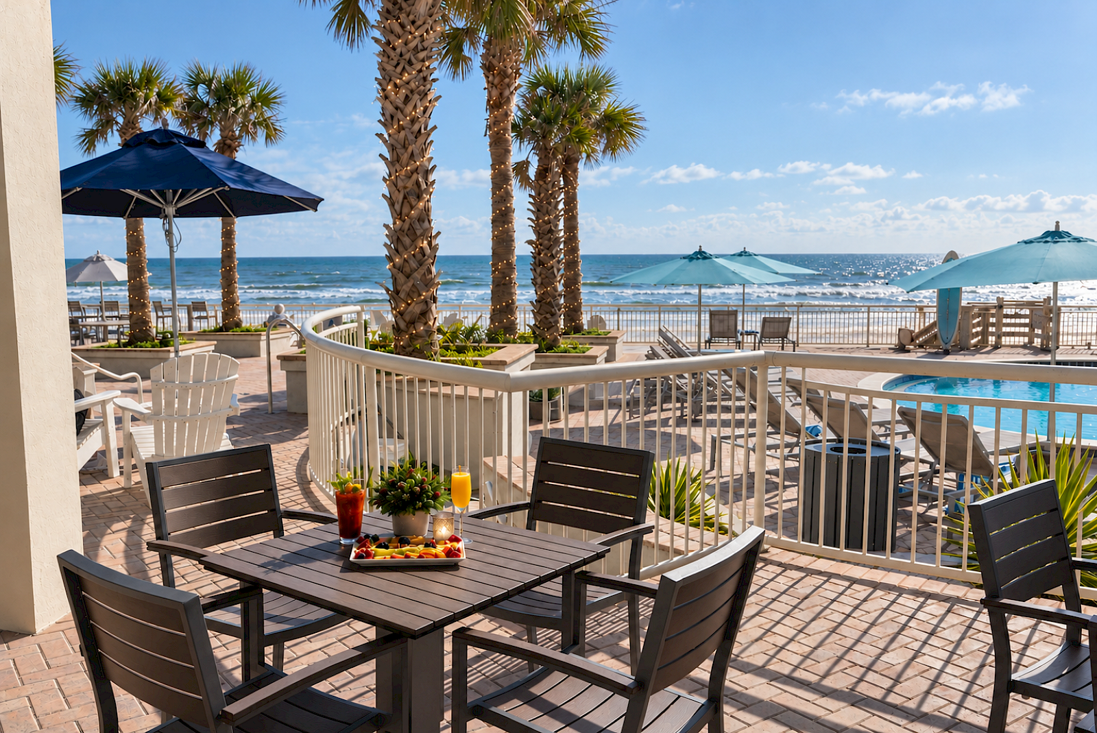 A sunny beachside patio with tables, chairs, umbrellas, palm trees, pool, and ocean views under a clear sky.