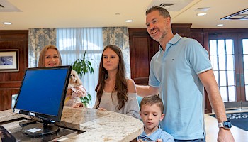 A man checks in a family at a hotel reception desk, with a computer, cozy decor, and welcoming atmosphere in the background.