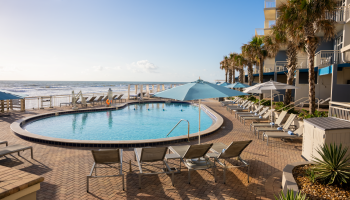 A resort pool area with lounge chairs, umbrellas, palm trees, and a view of the ocean on a sunny day.
