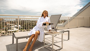 A woman in a white robe sits on a lounge chair on a rooftop, pouring red wine into a glass, enjoying the sunny view.