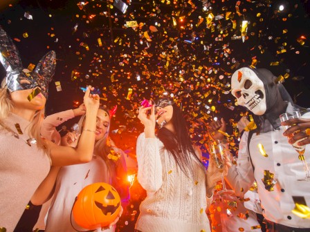 People celebrate Halloween with costumes, masks, confetti, and drinks, enjoying the festive spooky party atmosphere.