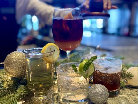 A bartender is preparing various colorful cocktails on a festive, decorated bar counter with holiday ornaments and greenery.