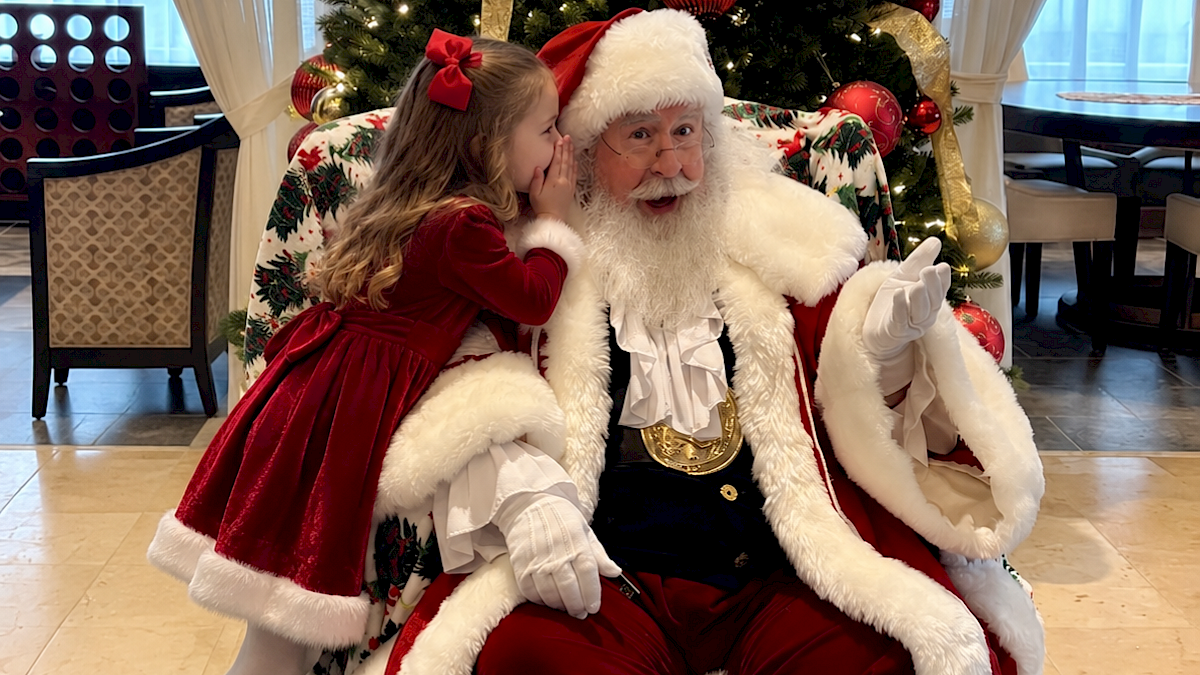 A young girl whispers to Santa Claus, who is sitting in front of a decorated Christmas tree, dressed in a red suit with a fluffy white trim.