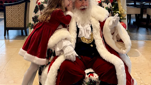 A young girl whispers to Santa Claus as he sits in a decorated armchair by a Christmas tree, sharing a festive moment.