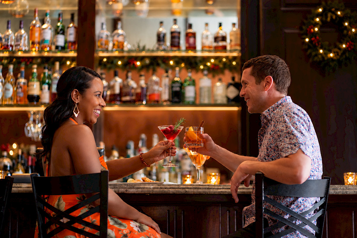 A man and woman enjoying cocktails at a festive bar, smiling and toasting each other during the holiday season.
