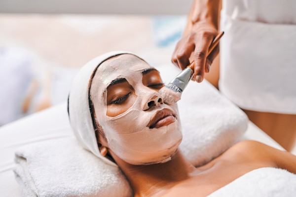A person is receiving a facial treatment, lying down with a white mask applied on their face, and a professional is using a brush to apply the mask.