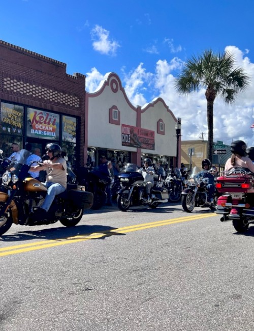 Bikers and motorcycles are parked outside a restaurant on a sunny day in a lively street scene.