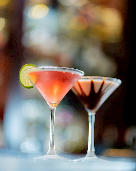 Two martini glasses with cocktails; one garnished with a lime slice and the other with chocolate, placed on a blurred background.