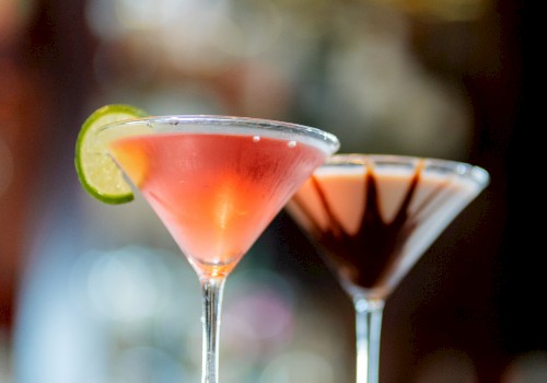 Two martini glasses with cocktails; one garnished with a lime slice and the other with chocolate, placed on a blurred background.