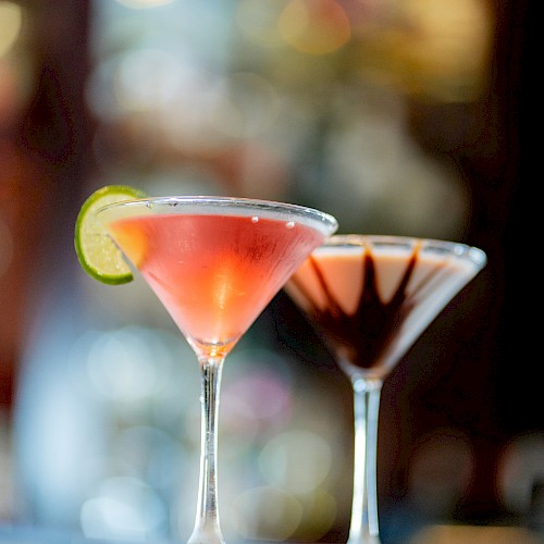 Two martini glasses with cocktails; one garnished with a lime slice and the other with chocolate, placed on a blurred background.