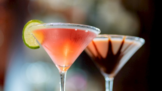 Two martini glasses with cocktails; one garnished with a lime slice and the other with chocolate, placed on a blurred background.