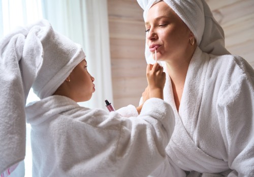A child in a bathrobe applies lip gloss to an adult also in a bathrobe and towel turban, both sharing a loving moment by a sunlit window.