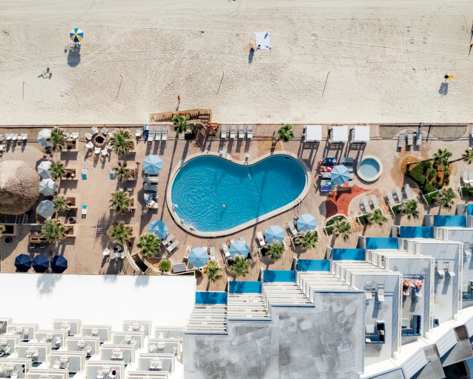 An aerial view of a beachside resort, featuring a heart-shaped pool with surrounding sun loungers, umbrellas, and people relaxing near the beach's edge.