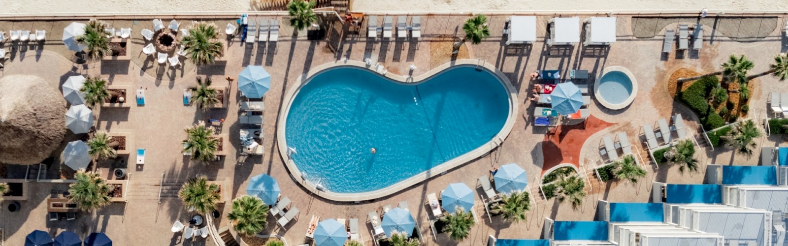 An aerial view of a beachside resort, featuring a heart-shaped pool with surrounding sun loungers, umbrellas, and people relaxing near the beach's edge.