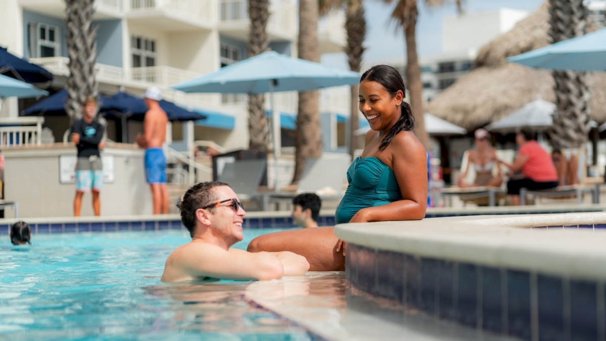 Two people are interacting by a pool. One person is in the pool, and the other person sits at the pool's edge. Others are in the background.