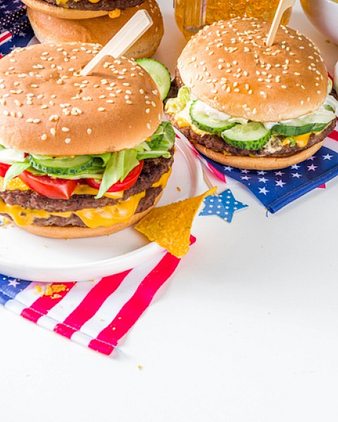 Two cheeseburgers with lettuce, tomato, and pickles on a patriotic-themed table, accompanied by fries and ketchup.
