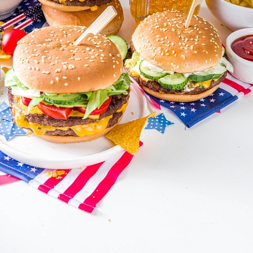 Two cheeseburgers with lettuce, tomato, and pickles on a patriotic-themed table, accompanied by fries and ketchup.