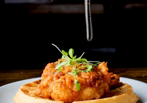 The image shows a plate of fried chicken and waffles being drizzled with syrup from a pitcher. The dish is garnished with greens.