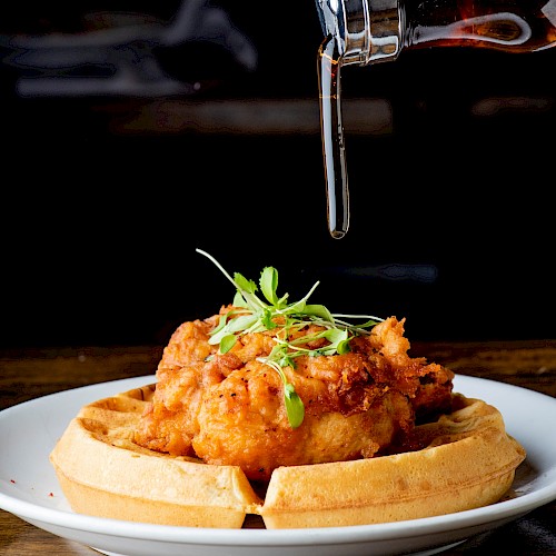 The image shows a plate of fried chicken and waffles being drizzled with syrup from a pitcher. The dish is garnished with greens.